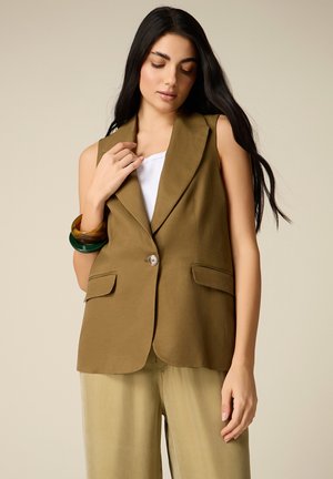 Donna con capelli lunghi e scuri che indossa un blazer senza maniche color oliva, un top bianco, pantaloni beige e bracciali verdi e marroni spessi, che posa su uno sfondo neutro.