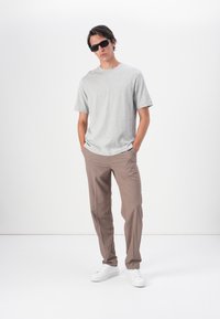 Tricou gri cu mânecă scurtă, pantaloni brown, adidași albi și ochelari de soare negri. Întregul outfit transmite o atmosferă casual și relaxată.