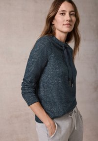 Tumman teal-värinen vetoketjullinen neule, joka on valmistettu teksturoidusta kankaasta, jossa on korkea kaulus, säädettävät narut ja pitkät hihat, yhdistettynä harmaisiin verkkareihin.