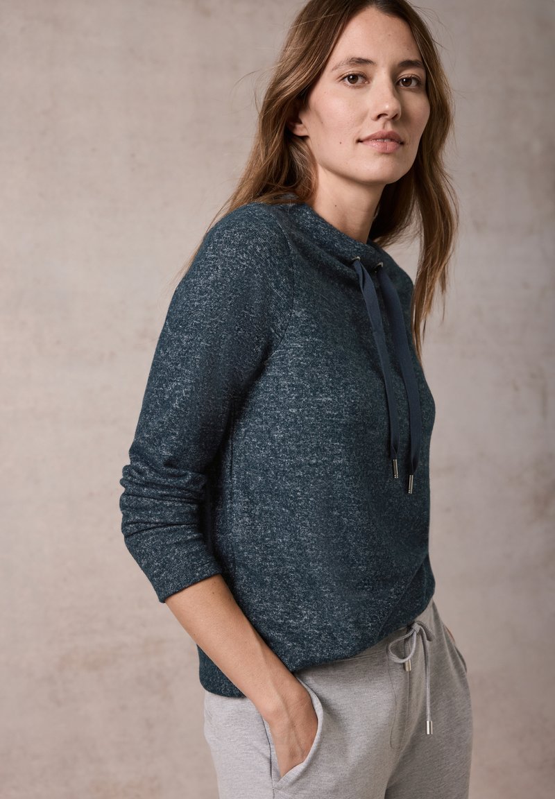 Tumman teal-värinen vetoketjullinen neule, joka on valmistettu teksturoidusta kankaasta, jossa on korkea kaulus, säädettävät narut ja pitkät hihat, yhdistettynä harmaisiin verkkareihin.