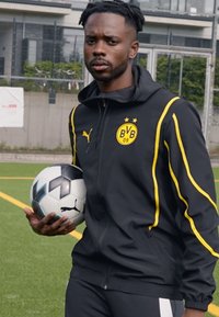Chaqueta deportiva negra con detalles amarillos y logo, pantalones deportivos, sosteniendo un balón de fútbol en blanco y negro. Campo de juego de fondo.