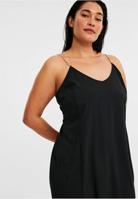 Schwarzes ärmelloses Kleid mit V-Ausschnitt, ausgestattet mit dünnen goldenen Ketten-Trägern und einer glatten, fließenden Textur. Schlichter und minimalistischer Stil.