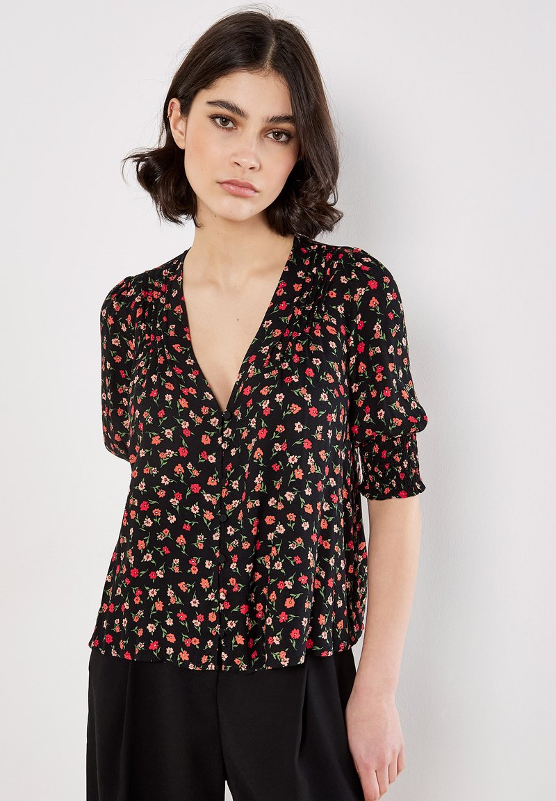 Zwarte bloemerige blouse met korte mouwen en een V-hals, versierd met kleine rode en roze bloemen, met geplooide details op de schouders en een knoopsluiting aan de voorkant.