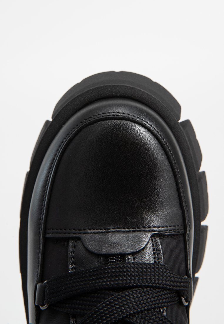 Schwarze Sneakers mit einer glänzenden Lederkappe und einem strukturierten Stoff-Obermaterial. Dicke Gummisohle mit geschichtetem Design. Die Schnürsenkel bestehen aus schwarzem gewebtem Material.