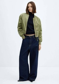 Frau in einer olivgrünen Bomberjacke, schwarzem Rollkragenpullover, weit geschnittenen, dunklen Jeans und schwarzen spitzen Schuhen, die vor einem weißen Hintergrund steht.