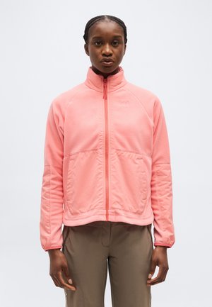 Femme aux cheveux tressés portant une veste polaire rose à fermeture éclair et un pantalon marron, debout devant un fond clair uni.