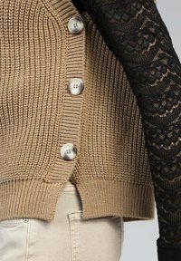 Cardigan en tricot beige avec un tissu texturé, un ourlet côtelé et trois grands boutons en argent. Une manche en dentelle noire est partiellement visible.