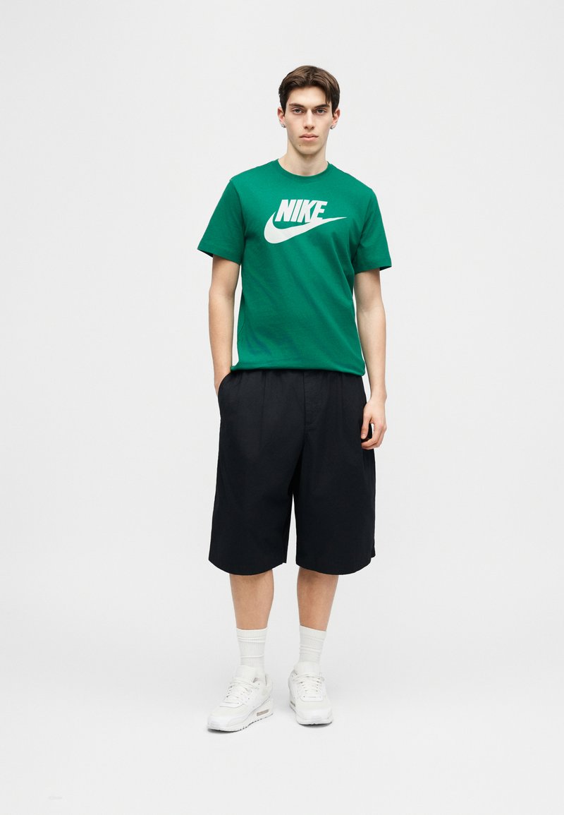 Junger Mann steht mit den Händen in den Taschen, trägt ein grünes Nike-T-Shirt, schwarze weite Shorts, weiße Socken und weiße Turnschuhe vor einfarbigem Hintergrund.