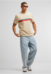 Camiseta de algodón beige con una franja horizontal roja, verde y negra; combinada con vaqueros anchos azul claro y zapatillas grises.