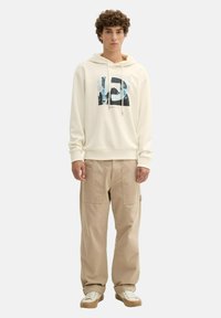 Sudadera con capucha crema con un diseño gráfico, combinada con pantalones cargo beige y zapatillas blancas con suela beige.