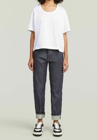 Camiseta blanca recortada con mangas cortas, combinada con jeans de mezclilla azul oscuro y doblados. Zapatillas negras y blancas completan el atuendo.