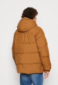Brun puderjakke med hætte, med horisontal quiltning og elastiske manchetter. Båret over blå jeans, hvilket får det teksturerede materiale til at skinne.