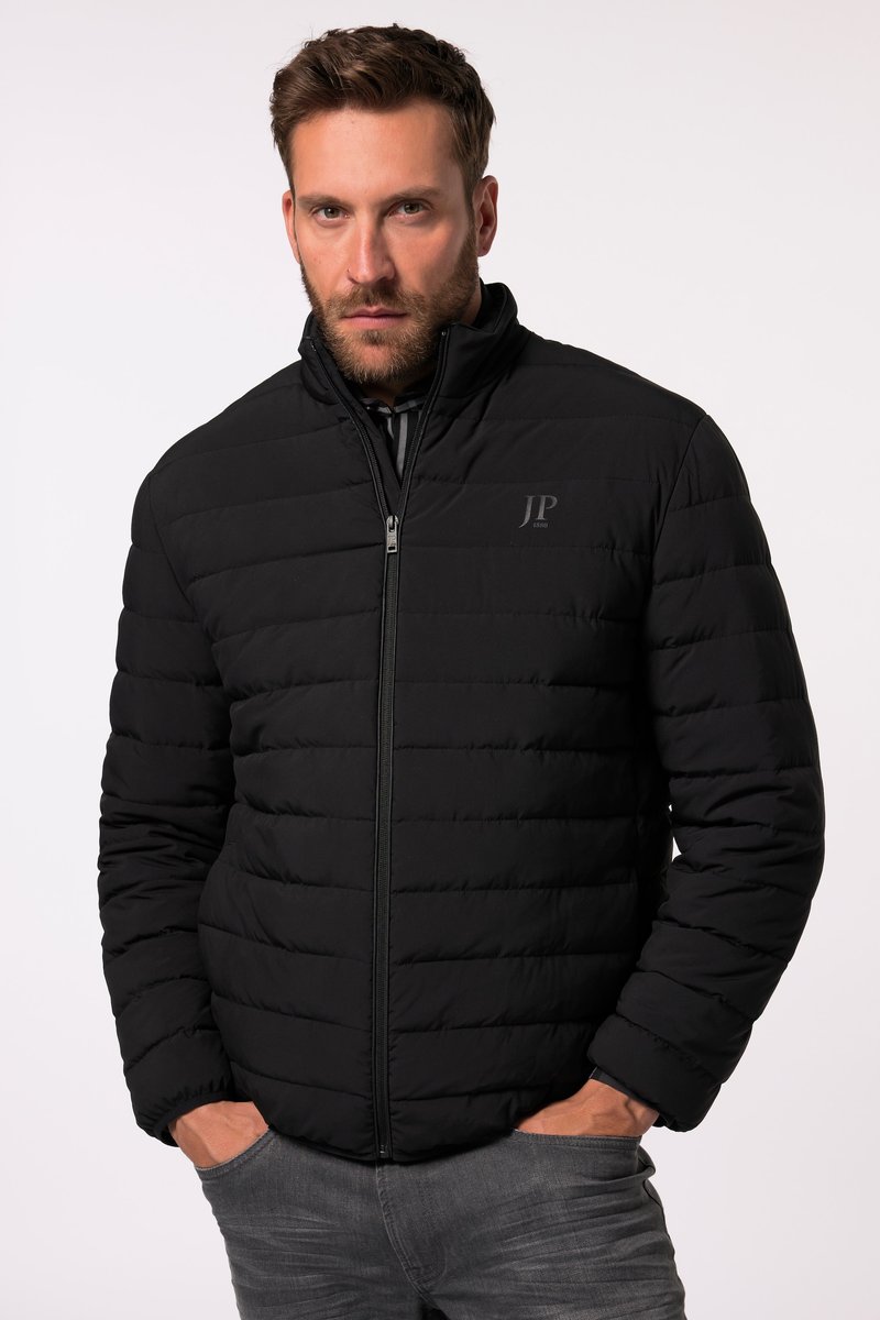 Mann trägt eine schwarze gesteppte Jacke mit den Händen in den Taschen und schaut nach vorne vor einem schlichten hellen Hintergrund.