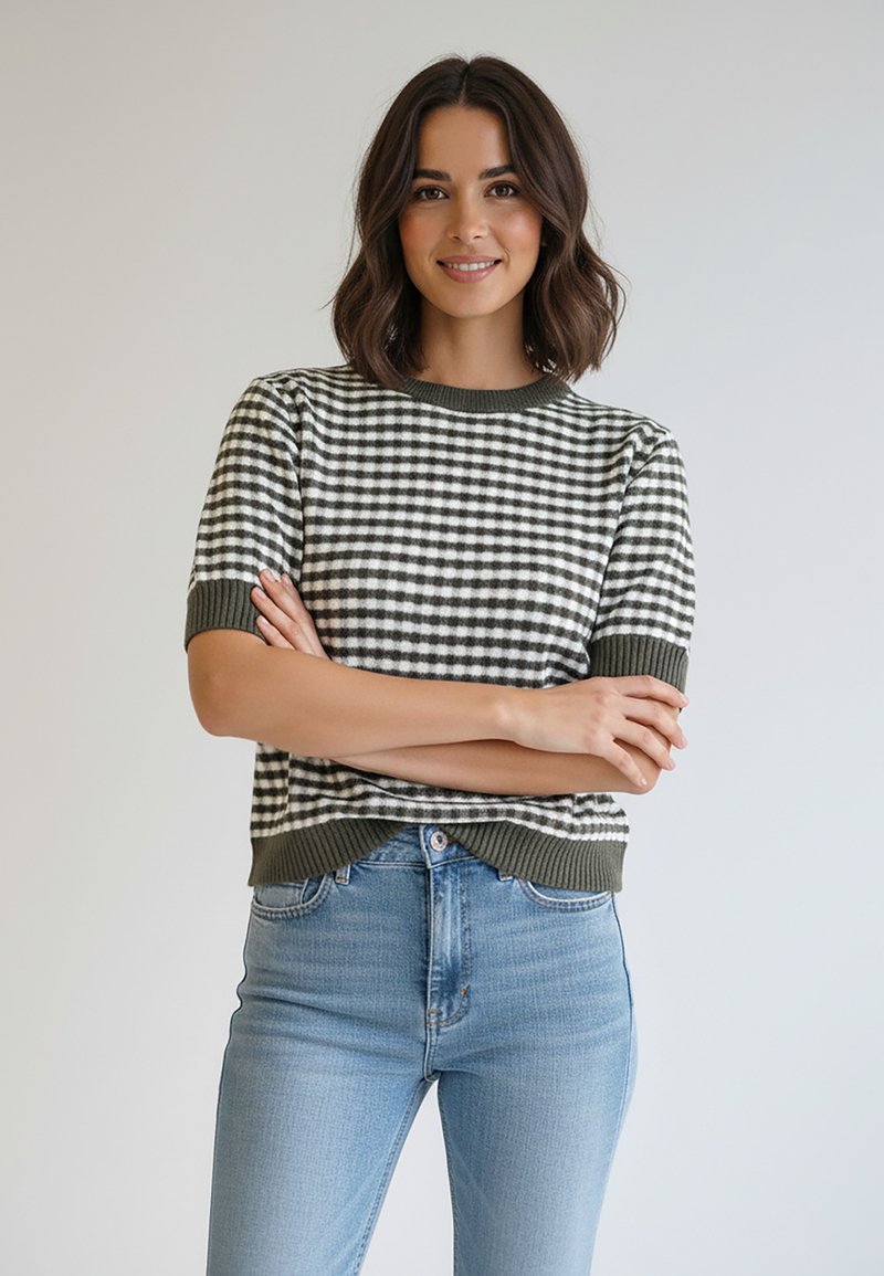 Sweter z krótkim rękawem w oliwkowo-białą kratkę. Ma prążkowany dekolt i dół, noszony z jasnoniebieskimi dżinsami.