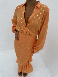 Oranges Kleid mit goldpunktiertem Muster, langen Ärmeln und gesmoktem Saum; kombiniert mit braunen Sandalen. Glatte Stofftextur.