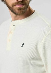 Krémový pletený svetr s dlouhým rukávem a knoflíkovým límcem typu henley, zdobený černou výšivkou postavy na hrudi. Texturovaná tkanina.