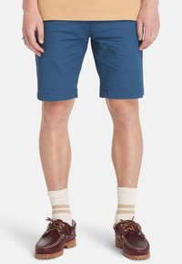 Pantaloni corti in cotone blu con design a gamba dritta, dotati di orli cuciti e tasche laterali, abbinati a scarpe marroni e calze a righe crema.