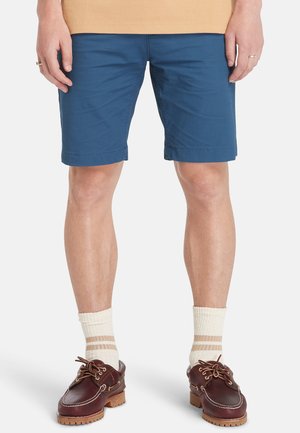 Blauwe katoenen shorts met een recht ontwerp, voorzien van gestikte zomen en zijzakken, gecombineerd metbruine schoenen en crèmekleurige gestreepte sokken.