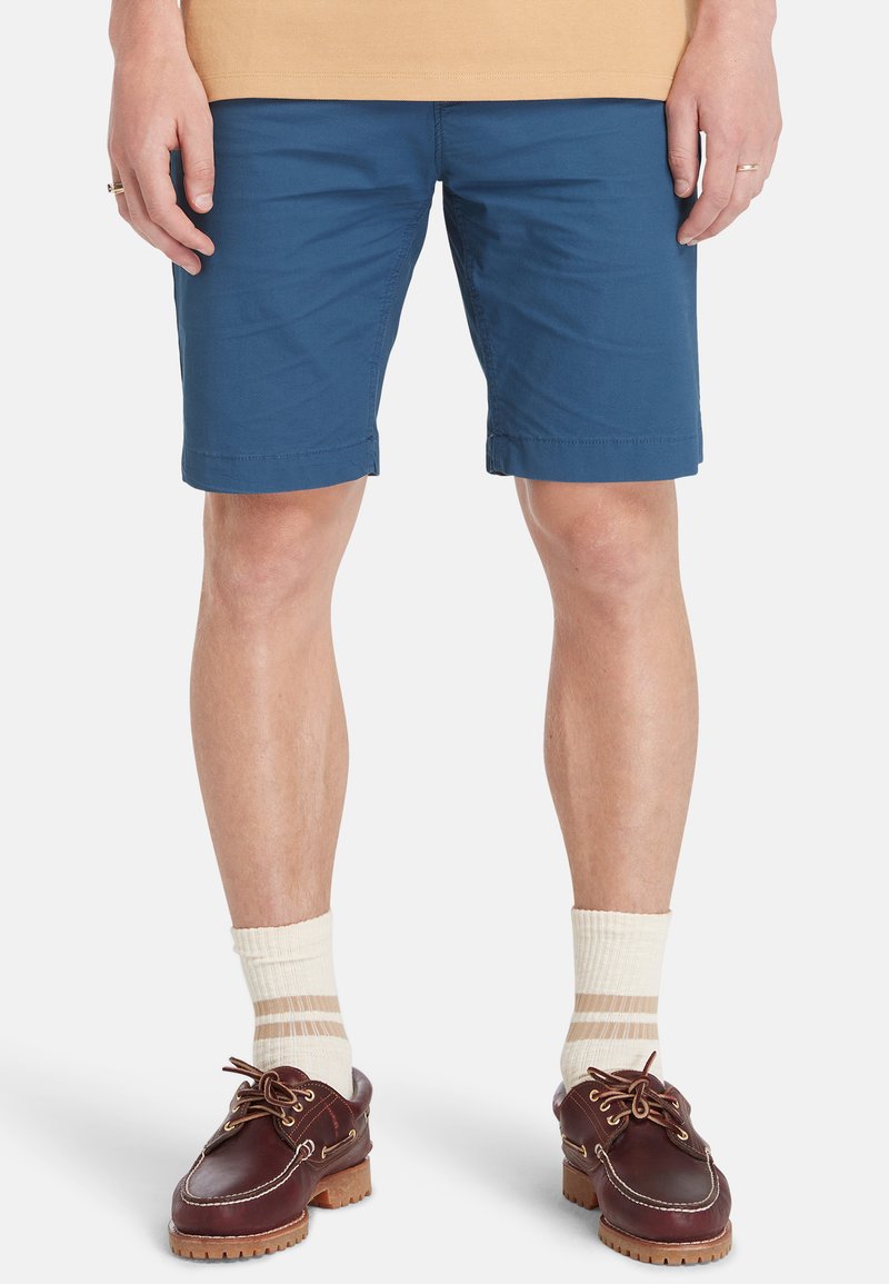 Pantaloni corti in cotone blu con design a gamba dritta, dotati di orli cuciti e tasche laterali, abbinati a scarpe marroni e calze a righe crema.