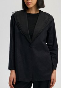 Černý blazer s texturovaným límcem, zapínání na jeden knoflík, dlouhé rukávy a mírně přiléhavý střih. Obsahuje čisté linie a minimální detaily.