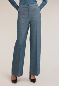 Wijde pijpenbroek in een gestructureerde blauwe stof, met een knoopsluiting en voorplooien. Gecombineerd met bordeauxrode hoge hakken.