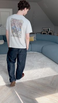 T-shirt cinza com um gráfico vintage da Levi Strauss, emparelhado com jeans azul escuro e mocassins castanhos. Sofá azul claro curvado e tapete branco visíveis.