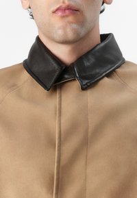 Veste en daim marron avec un col en cuir noir, présentant un design structuré et des coutures visibles sur le devant.