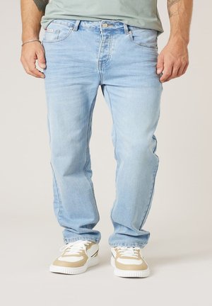 Man draagt lichtblauwe spijkerbroek en beige-witte sneakers, staand met ontspannen handen langs het lichaam op een neutrale achtergrond.