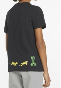 Persona che indossa una maglietta nera con ghepardi gialli pixelati e un personaggio verde squadrato sul retro in basso, in piedi contro uno sfondo bianco.