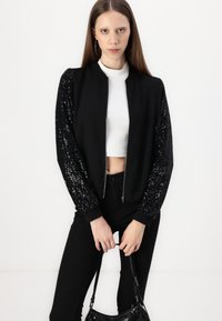 Schwarze Bomberjacke mit Paillettenärmeln, Reißverschluss vorne, Rippbündchen und kurzer Passform; getragen über einem weißen Oberteil und kombiniert mit schwarzen Hosen.