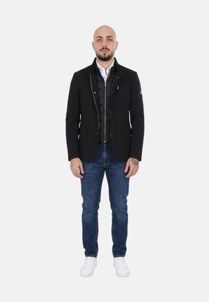 Uomo in piedi che indossa un blazer nero sopra una giacca nera con cerniera, jeans blu e sneakers bianche su uno sfondo uniforme.