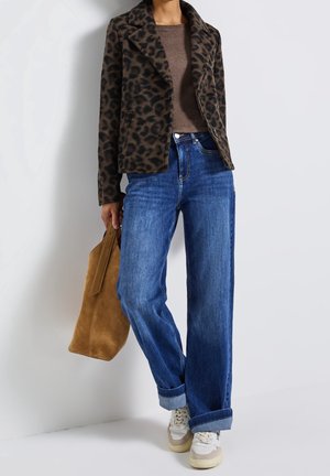 Person trägt eine Jacke mit Leopardenmuster, braunes Oberteil, hochgekrempelte blaue Jeans, weiße Turnschuhe und hält eine große braune Wildledertasche.