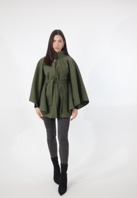 Grøn kappe i uldblanding med høj krave og bælte, med vide ærmer og afslappet pasform. Kombineret med slanke, mørkegrå jeans og sorte ankelstøvler.