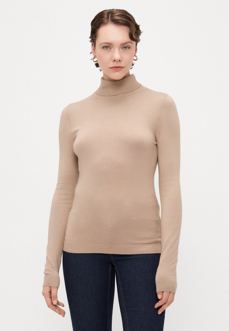 Beige turtleneck sweater with long sleeves, ribbed hem, and a smooth texture. Worn with dark blue jeans, featuring fitted design.