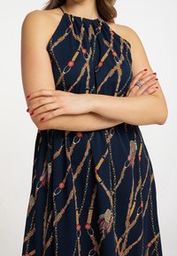 Mörkblå халтерklänning med samlad halsringning och ett mönster av kedjor, knappar och akcentfärger i röd, guld och orange. Mjuk tygtextur.