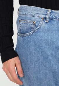 Jasnoniebieskie dżinsy w klasycznym kroju, z przednimi kieszeniami i metalowymi akcentami. Noszone z czarnym swetrem w prążki.