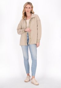 Gepolsterte beige Jacke mit kragenförmigem Ausschnitt, ausgestattet mit großen Knöpfen, kombiniert mit hellblauen Jeans und beigen Sneakers. Glatte Stoffstruktur.