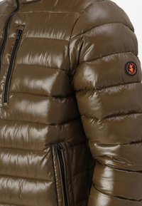 Veste matelassée vert olive à finition brillante, avec un quilting horizontal, une fermeture éclair à l'avant et un patch logo orange sur la manche.