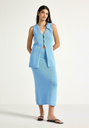 Ung kvinde iført lyseblå ærmeløs vest med knapper og matchende langt skørt samt sorte sandaler, stående på hvid baggrund.