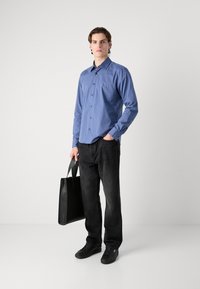 Cămașă albastră cu mânecă lungă și nasturi, blugi negri și adidași negri. Ținând o geantă mare neagră de umăr. Design simplu cu o croială lejeră.