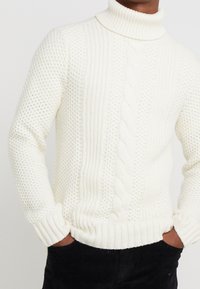 Maglione bianco a collo alto in maglia con un motivo testurizzato, caratterizzato da dettagli a treccia e polsini e orlo a coste. Indossato sopra pantaloni neri.