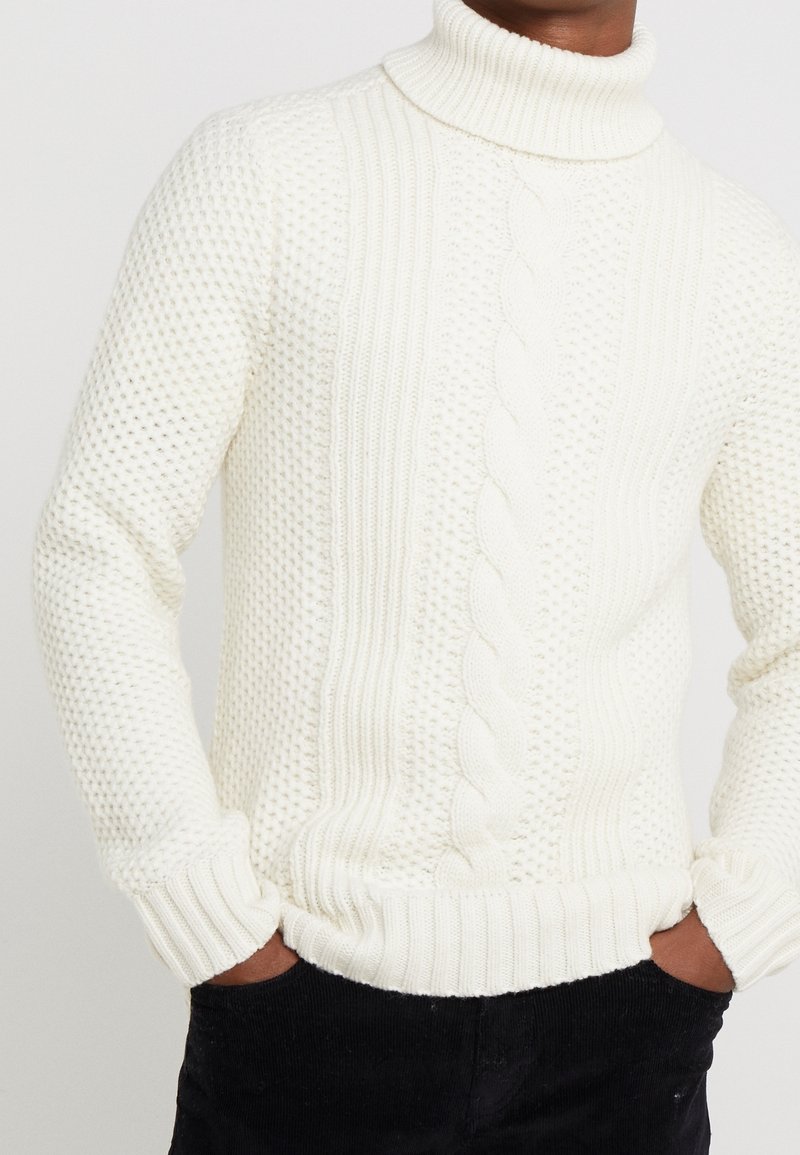 Maglione bianco a collo alto in maglia con un motivo testurizzato, caratterizzato da dettagli a treccia e polsini e orlo a coste. Indossato sopra pantaloni neri.