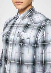 Camicia a quadri blu e nera con bottoni e due tasche, caratterizzata da un collo con risvolto e cuciture a contrasto. Il tessuto ha una texture liscia.