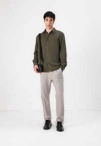 Camisa verde oliva de botones, pantalones grises y zapatos negros. Modelo sostiene un pequeño bolso negro, de pie contra un fondo blanco liso.