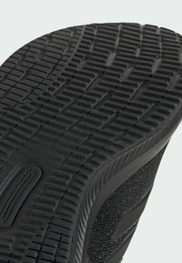 Semelle de sneaker noire avec une surface en caoutchouc texturée, présentant un motif en grille et des crampons supplémentaires pour l'adhérence. Sur le côté, on peut voir un tissu tissé.