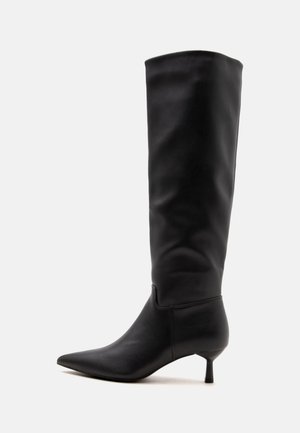 Botte en cuir noire montante jusqu'au genou, à bout pointu et talon kitten fin, sur fond clair uni.