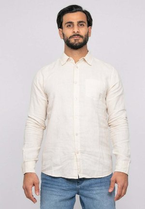 Homme aux cheveux foncés et barbe, portant une chemise en lin crème boutonnée et un jean bleu, debout devant un fond gris clair.