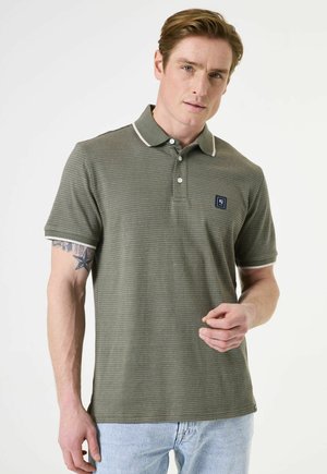 Khakifarbenes Polohemd mit strukturiertem Design, weißen Kragen und Ärmeln sowie einem kleinen dunkelblauen Logo-Patch auf der Brust; kombiniert mit hellen Jeans.