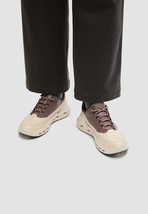 Låga, vida svarta byxor som bärs med tvåfärgade, tjocka sneakers i beige och brunt, med tjocka sulor och mönstrade skosnören.