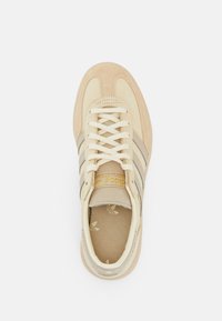 Zapatilla deportiva beige con parte superior de ante y tela, tres franjas horizontales beige, cordones blancos y logotipo dorado en la lengüeta.
